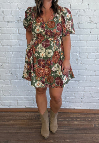 A woman modeling a short-sleeve floral mini dress with a V-neck, standing in front of a white brick wall and dark wood floor.