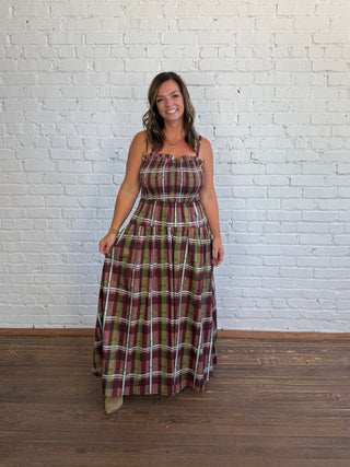 A woman modeling a sleeveless, floor-length plaid maxi dress with thin straps, standing against a white painted brick wall on a wooden floor.