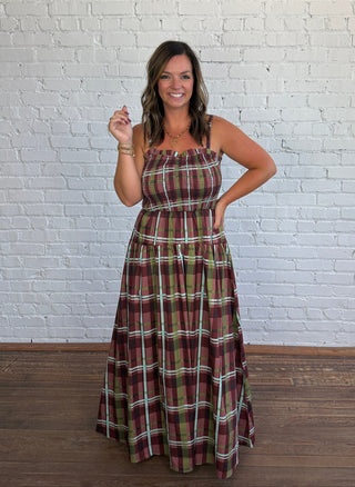 A woman modeling a thin strap plaid maxi dress with a smocked bodice, standing against a white brick wall and wooden floor.