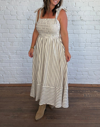 A woman wearing a sleeveless cream and taupe vertically striped maxi dress with tie shoulder straps, standing in front of a white painted brick wall on a dark wood floor.