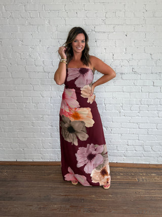 A woman wearing a strapless maxi dress with a large floral print in burgundy, pink, purple and beige, standing against a white brick wall and wooden floor.