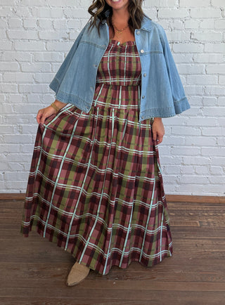 A woman stands in front of a white brick wall wearing a long burgundy, olive, and blue plaid dress with a light blue denim jacket draped over her shoulders and tan pointed shoes.