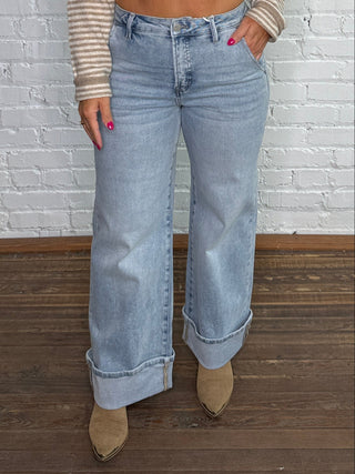 A woman stands against a white brick wall wearing a beige and white striped long-sleeve button-up shirt, light-wash wide-leg jeans, and tan pointed-toe heels.
