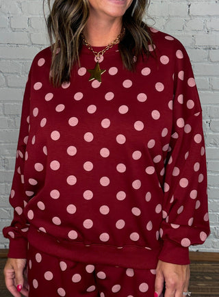 A woman modeling a matching burgundy and pink polka dot two-piece set standing against a white brick wall and wooden floor.