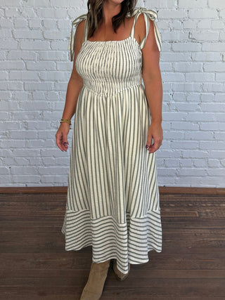 A woman modeling a sleeveless midi dress with vertical cream and olive green stripes, featuring tie-shoulder straps and a smocked bodice, standing against a white brick wall and dark wood floor.