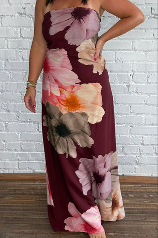 A woman wearing a strapless maxi dress with a large floral print in burgundy, pink, purple and beige, standing against a white brick wall and wooden floor.