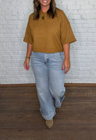 A woman stands in front of a white brick wall wearing a mustard brown short-sleeve top, light blue jeans, and tan boots.