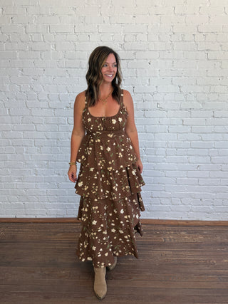 A woman modeling a brown tiered maxi dress with a white floral pattern, standing in front of a white brick wall on a wooden floor.