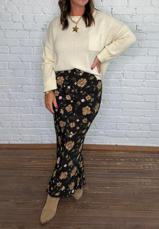 A woman posing in a cream knit sweater and black floral maxi skirt with beige boots, standing against a white brick wall and wood floor.