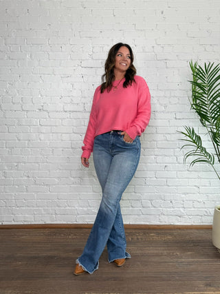 Woman in a pink sweatshirt and blue jeans standing against a white brick wall with a plant to her right.