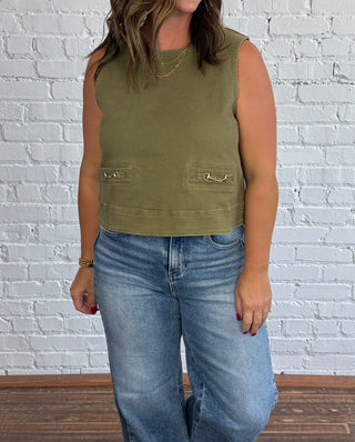 A woman stands in a room with a white brick wall and dark wood floor, wearing a sleeveless olive green top with gold equestrian horse bit faux pocket detail and wide-leg blue jeans.