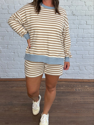 A woman modeling a matching taupe and cream striped lounge set with light blue trim, consisting of a long-sleeve sweatshirt and shorts, standing against a white brick wall and wood floor.