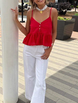 Woman in a red top and white pants standing on a paved area with trees and buildings in the background.