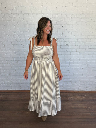 A woman modeling a sleeveless, square-neck maxi dress with thin shoulder ties and vertical stripes, standing against a white brick wall and dark wood floor.