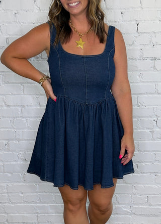 A woman modeling a dark blue denim-like sleeveless dress with a gathered waist and flared skirt.
