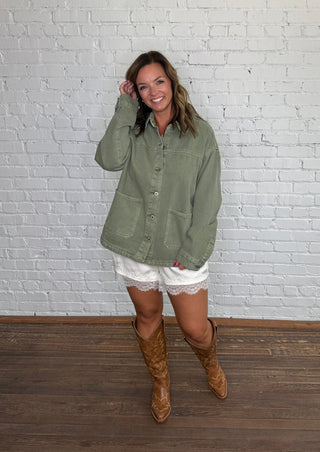 A woman stands in front of a white brick wall wearing an olive green button-up jacket, white shorts, and brown cowboy boots.