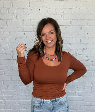 A woman posing in a rust long-sleeve scoop-neck top, light-wash blue jeans, and brown shoes, standing against a white brick wall and wooden floor.