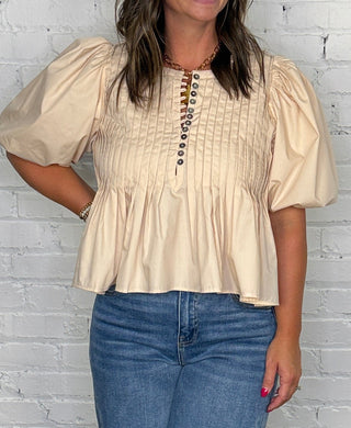 A woman modeling a cream-colored short-sleeve blouse with puff sleeves and a button-up front, paired with wide-leg blue jeans, standing against a white brick wall and wooden floor.