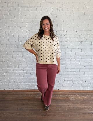A woman stands in a studio with a white brick wall and wood floor, wearing a cream knit sweater with brown polka dots and dusty rose pants.