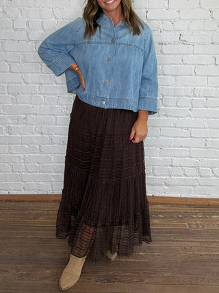 A woman stands against a white brick wall wearing a light blue cropped denim jacket, a dark brown lace tiered midi skirt, and tan ankle boots.