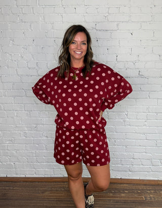 A woman modeling a matching burgundy and pink polka dot two-piece set standing against a white brick wall and wooden floor.