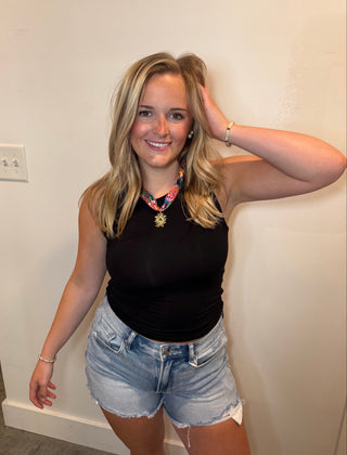 Woman wearing a black tank top and denim shorts standing against a plain wall.