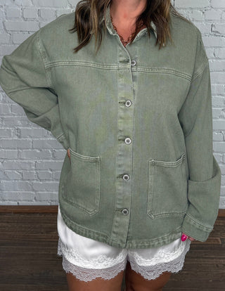 A woman wearing an oversized sage green button-up shirt jacket, white lace-trimmed shorts, and brown knee-high boots, standing against a white brick wall.