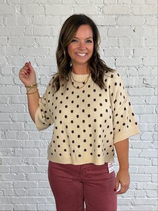 A woman stands in a studio with a white brick wall and wood floor, wearing a cream knit sweater with brown polka dots and dusty rose pants.