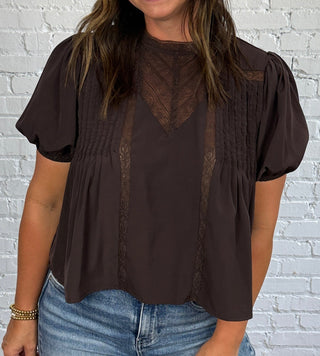A woman wearing a dark brown short-sleeve blouse with a sheer mesh front panel and pleated details, paired with medium wash blue jeans, standing in front of a white brick wall.