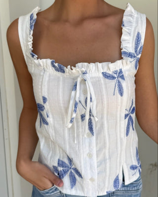 Woman wearing a white sleeveless top with blue floral patterns and denim shorts.