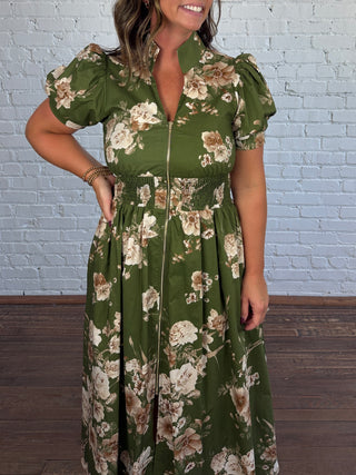 A woman wearing an olive green midi dress with a beige and white floral print, puff sleeves, and a smocked waist, standing against a white brick wall.