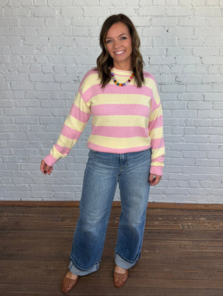 A woman wearing a pink and yellow striped knit sweater, blue jeans, and brown shoes, posing against a white brick wall and dark wood floor.