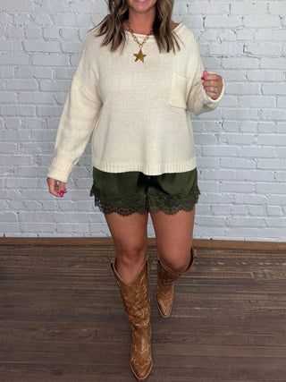 A woman modeling a cream knit sweater, olive green lace shorts, and brown cowboy boots, standing against a white brick wall and dark wood floor.