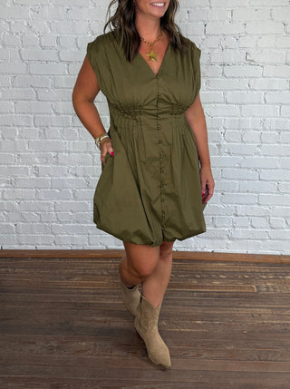 A woman modeling an olive green sleeveless bubble-skirt, mini dress with a gathered waist, paired with beige ankle boots, standing against a white brick wall and dark wood floor.