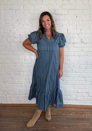 A woman wearing a blue and green vertically striped midi dress with puff sleeves and beige ankle boots, standing against a white brick wall on a wooden floor.