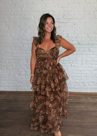 A woman modeling a brown floral tiered maxi dress with ruffled shoulder straps, standing against a white brick wall and dark wood floor.