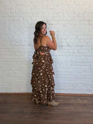 A woman wearing a brown tiered maxi dress with a beige floral print, standing in front of a white brick wall on a wooden floor.
