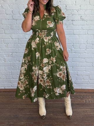 A woman models a green midi dress with a beige and white floral pattern, featuring puff sleeves, a smocked waist, and a front zipper. She is wearing cream-colored pointed-toe boots and standing in front of a white brick wall and dark wood floor.