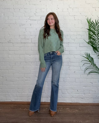 Person wearing a green sweater and blue jeans standing on a white brick floor with a plant in the corner.