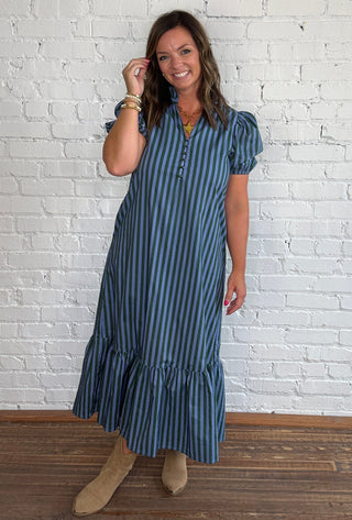 A woman wearing a blue and green vertically striped midi dress with puff sleeves and beige ankle boots, standing against a white brick wall on a wooden floor.