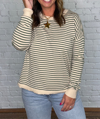 A woman stands in front of a white painted brick wall wearing a cream and dark green striped long-sleeve knit top with light blue jeans.