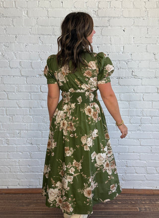 A woman viewed from behind wearing a green midi dress with a beige and white floral pattern, standing on a dark wood floor against a white brick wall.