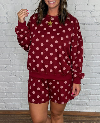A woman modeling a matching burgundy and pink polka dot two-piece set standing against a white brick wall and wooden floor.