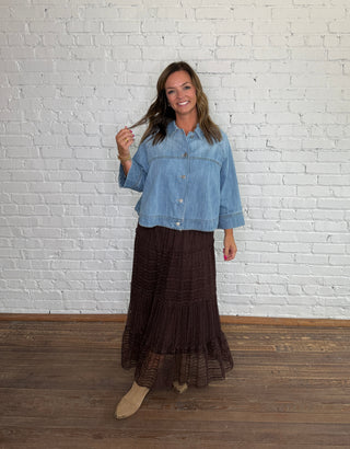 A woman stands in front of a white brick wall wearing a cropped denim top and a dark brown tiered lace midi skirt, paired with taupe ankle boots.