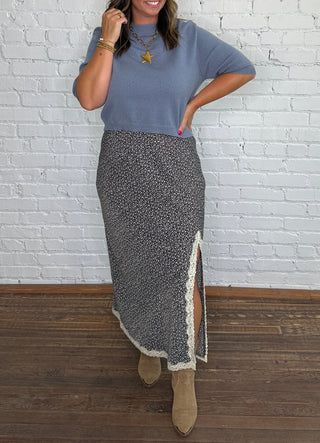A woman wearing a dusty blue short-sleeve top, a brown and blue floral midi skirt with a cream lace side slit, and tan ankle boots.