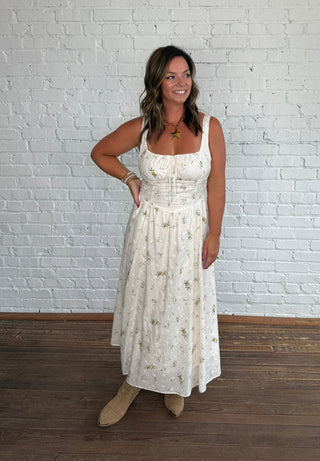 A woman modeling a spaghetti strap cream maxi dress with a delicate floral pattern, standing against a white brick wall on a dark wood floor.