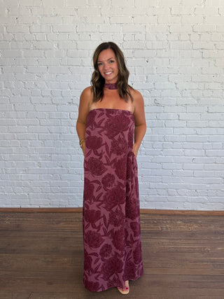 A woman modeling a strapless maxi dress with a tonal floral pattern in deep burgundy and plum, standing against a white brick wall.