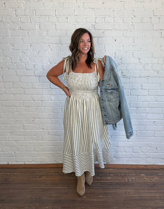 A woman stands in front of a white brick wall wearing a cream and olive vertically striped sundress with a smocked bodice and ruffled hem, holding a light-wash denim jacket over her arm. She is wearing tan ankle boots.