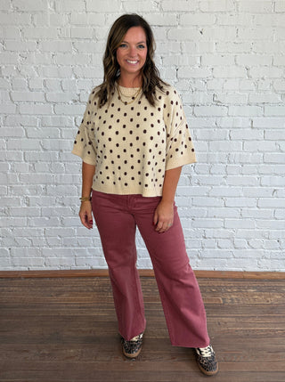 A woman stands in a studio with a white brick wall and wood floor, wearing a cream knit sweater with brown polka dots and dusty rose pants.
