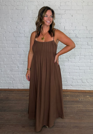 A woman modeling a brown sleeveless midi dress with a pleated bodice and flowing skirt.
